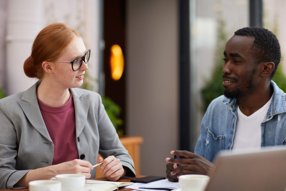 Multiethnic young business colleagues talking to each other while sitting at the table with documents and laptop at meeting in cafe